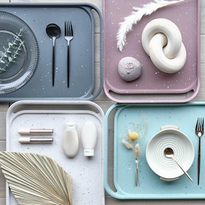 May include: Four different colored trays with various items on them. The trays are made of a speckled material and have handles on the sides. The items on the trays include a glass plate, a fork, a spoon, a ring, a wooden knot, a palm leaf, two tubes of lip gloss, two bottles of lotion, a small bouquet of flowers, a bowl, a spoon, and a fork.