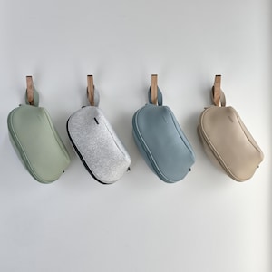 May include: Four small, soft-sided bags in pastel colors hang from wooden pegs on a white wall. The bags are in shades of green, gray, blue, and beige. Each bag has a small handle and a zipper closure.