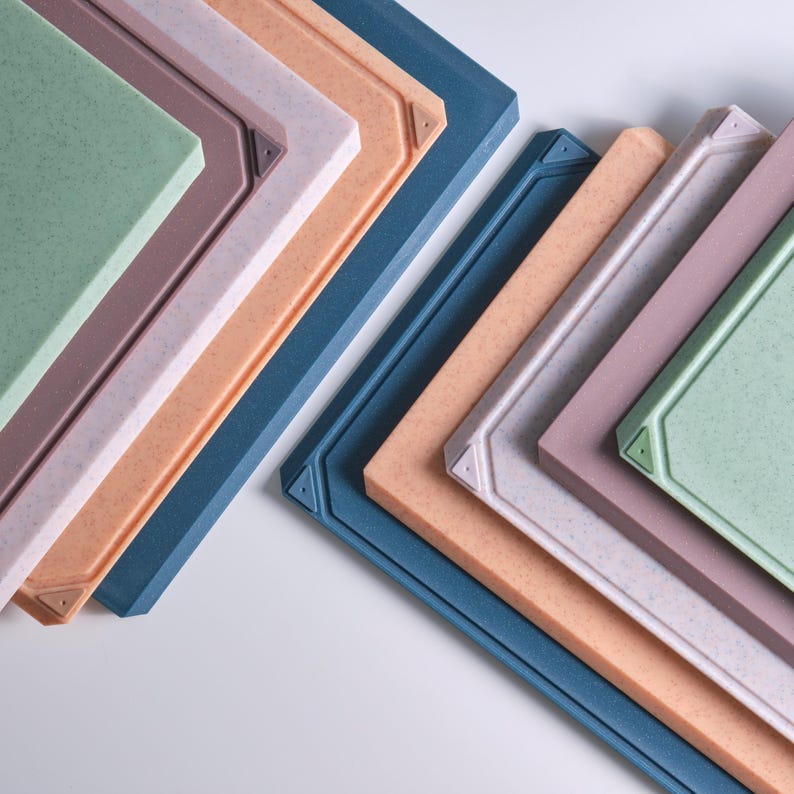 May include: An assortment of colorful cutting boards in shades of blue, orange, pink, green, and brown. Each board has a textured surface and a decorative border with a corner design. The boards are arranged in a diagonal pattern.