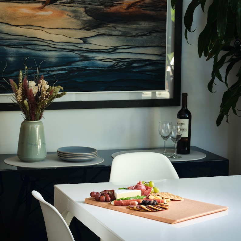 May include: A dining scene with a white table set for a meal. A charcuterie board with grapes, cheese, and crackers sits on the table. A vase of flowers, wine glasses, and a bottle of wine are also present. A large painting hangs on the wall.