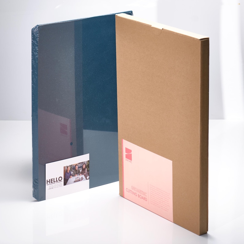 May include: Two rectangular boxes, one navy blue and one brown, stand side-by-side. The blue box is wrapped in clear plastic. The brown box has a pink label that reads "INNOVATIVE CUTTING BOARD". A white label on the blue box says "HELLO".