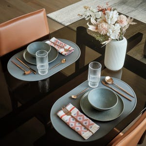 May include: A dining table setting with two place settings. Each setting includes a blue-grey oval placemat, a bowl, a plate, a glass, a spoon, a fork, and a patterned napkin. A white vase with flowers is in the center.