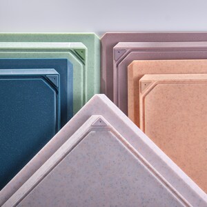 May include: A collection of colorful, square cutting boards in shades of blue, green, pink, and peach. Each board has a textured surface and a geometric design with a small hole in the corner. The boards are stacked, showcasing their varying sizes.