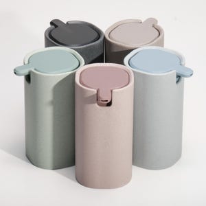 May include: Five cylindrical wastebaskets in various pastel colors: gray, pink, blue, green, and taupe. Each has a matching lid with a handle. The wastebaskets are arranged in a circular pattern against a white background.