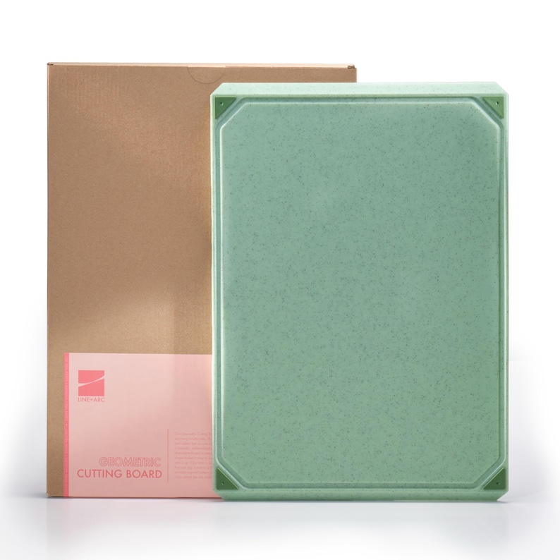 May include: A rectangular, light green cutting board with a speckled texture, next to a brown cardboard box. The cutting board has a slightly raised edge and corner accents. The box has a pink label with the text "GNOMONIC CUTTING BOARD".