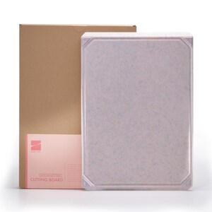 May include: A light pink cutting board with a speckled pattern and a geometric design. The cutting board is rectangular with a slightly raised edge. A brown cardboard box and a pink card with the text "GEOMETRIC CUTTING BOARD" are in the background.