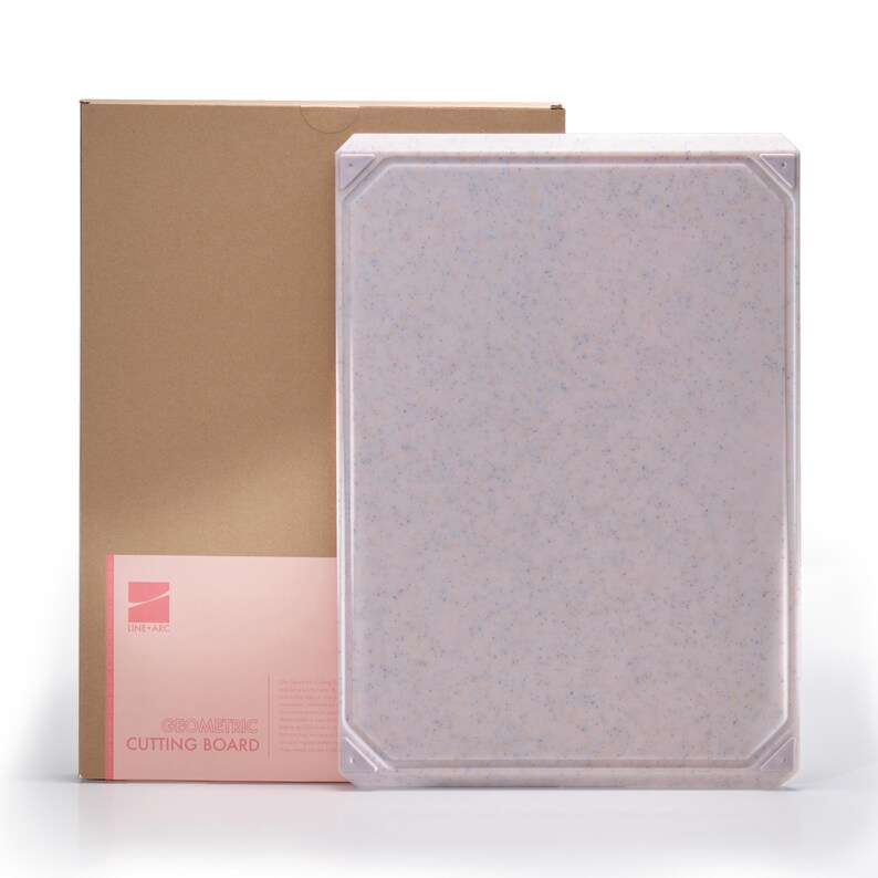May include: A light pink cutting board with a speckled pattern and a geometric design. The cutting board is rectangular with a slightly raised edge. A brown cardboard box and a pink card with the text "GEOMETRIC CUTTING BOARD" are in the background.