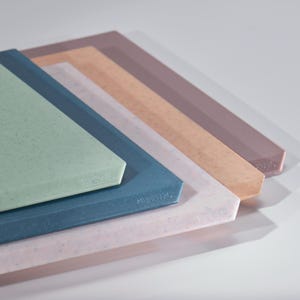 May include: A stack of rectangular cutting boards in various colors, including blue, green, pink, peach, and brown. Each board has a speckled texture and a beveled edge. The boards are arranged at an angle, showcasing their different hues.