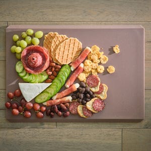 May include: A charcuterie board with a variety of snacks. Includes green and red grapes, salami, cucumber slices, cheese, crackers, nuts, olives, and waffle cookies. The board is a rectangular shape and is a muted purple color.