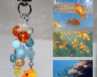 May include: A keychain featuring a silver-tone metal clasp and ring, adorned with a cluster of multi-colored beads in orange, blue, yellow, and clear glass, and a small orange goldfish-shaped charm hanging from the bottom.