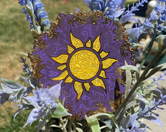 May include: A purple and gold enamel pin featuring a stylized yellow sun design. The pin has a decorative border and is set against a backdrop of lavender-colored flowers in a white pot. The sun design is intricate and detailed.