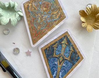 May include: Two square stickers with intricate designs. One features an owl in shades of orange, gold, and blue, while the other showcases a blue and gold aquatic scene. Both have a gold border and are on a white surface.