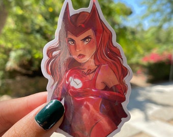 May include: A sticker featuring a red-clad character with a stern expression, long red hair, and a distinctive headpiece. The artwork has a comic book style with a glossy finish, set against a blurred green and blue background.