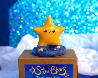 May include: A yellow star figurine with a smiling face, set on a blue base with star accents. The figurine sits atop a brown box with a blue and gold sticker that reads "StarBeans". The background is a sparkly blue.