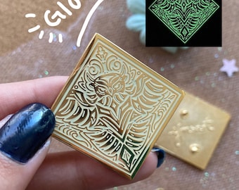 May include: Square gold-colored enamel pin with an owl design. The pin has a detailed, symmetrical pattern with a cream-colored background. The word "GLOW" is written in white. A smaller image shows the pin glowing green in the dark.