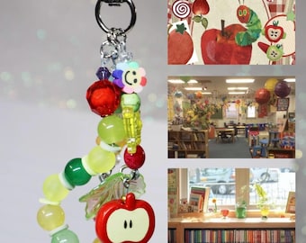 May include: A beaded keychain featuring a silver-tone lobster clasp, a small flower charm with a smiley face, a red apple charm, and various translucent green, yellow, and red beads. The design includes a small gold-tone star charm at the end.