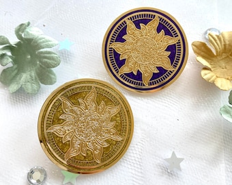 May include: Two gold-colored, circular pins with a sunburst design. One pin has a dark purple background, while the other has a gold background. The pins are embellished with intricate details and are set against a white background with decorative elements.