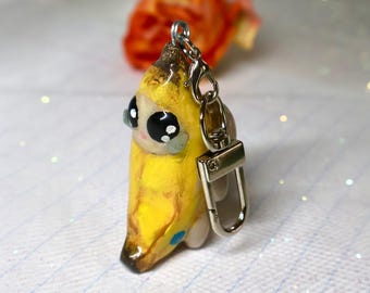 May include: A yellow banana-shaped keychain featuring a glossy finish, large black eyes with white highlights, and a brown stem tip. The figure is attached to a silver-toned metal lobster clasp and a split ring, creating a functional accessory.