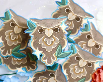 May include: A collection of die-cut stickers featuring a brown owl with large dark eyes, a light-colored beak, and intricate feather patterns. Each owl is accented with small green leaves and four-pointed white stars, outlined in a bright blue border.
