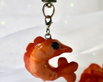May include: A glossy orange shrimp-shaped charm attached to a silver-toned metal lobster clasp keychain. The shrimp features a curved body, a prominent black eye, and a segmented tail, with a smooth, reflective finish throughout the piece.