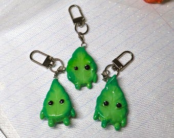 May include: Three green leaf-shaped keychains with small black eyes and tiny limbs. Each charm is attached to a silver-colored metal lobster clasp and ring. The leaf figures have a smooth, glossy finish and a vibrant, multi-tonal green color.