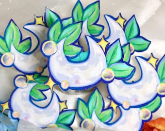 May include: A collection of crescent moon stickers featuring a light purple moon shape accented with vibrant green leaves, small yellow four-pointed stars, and translucent circular bubbles, all outlined in a bold dark blue border.