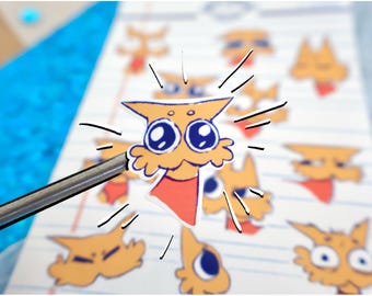 May include: A sheet of cartoon cat stickers in various poses. The stickers are orange with blue eyes and red accents. A metal tool is touching one sticker, which has a jagged edge and black lines radiating from it. The background is lined paper.