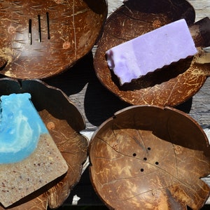 May include: Four dark brown coconut shell bowls shaped like leaves, two of which contain rectangular bars of soap. One soap bar is solid light purple, while the other features a blue top layer and a textured, speckled tan bottom layer.