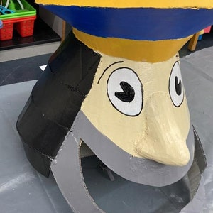 May include: A large cardboard cutout of a cartoon face with a yellow hat, blue brim, black hair, white eyes, and a gray beard. The face is painted with a light brown skin tone and has a large, gray, open mouth.