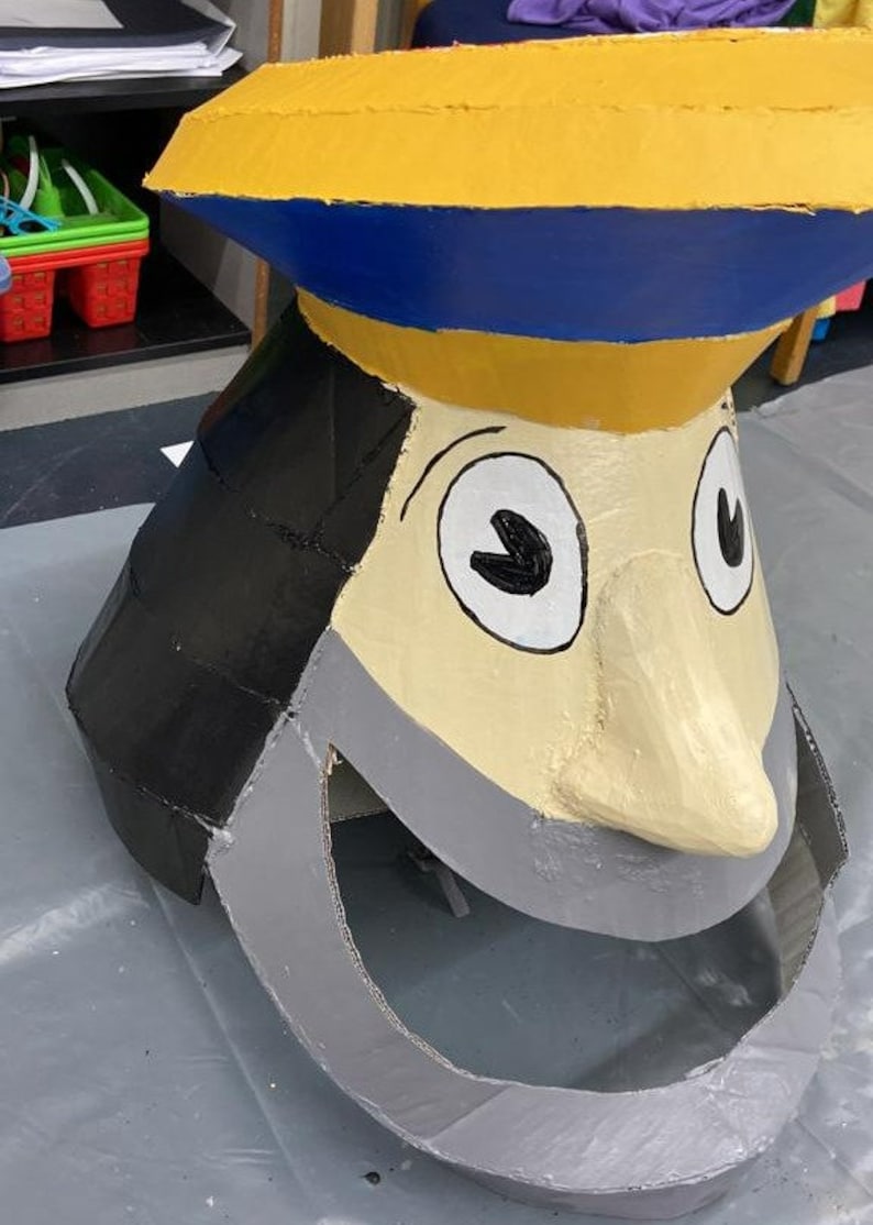 May include: A large cardboard cutout of a cartoon face with a yellow hat, blue brim, black hair, white eyes, and a gray beard. The face is painted with a light brown skin tone and has a large, gray, open mouth.