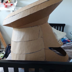 May include: A large cardboard sculpture of a head, possibly a character from a cartoon or video game. The head is made of multiple layers of cardboard, and the details are sculpted with tape and glue. The head is painted white in some areas.