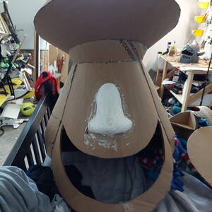 May include: A cardboard head sculpture with a white paper mache nose. The sculpture is unfinished and has a large opening for the face.