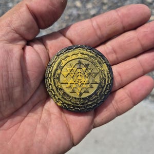 May include: A circular black and gold medallion featuring a detailed Sri Yantra geometric pattern. The design consists of a central star composed of interlocking triangles surrounded by concentric circular borders with intricate petal-like motifs.