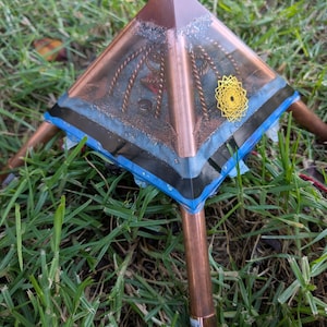 Op de afbeelding: Een koperkleurige piramide met een transparante harslaag, met een gouden geometrisch ontwerp en ingebed koperdraad. De piramide wordt ondersteund door vier koperen buizen en ligt op groen gras.