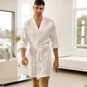 May include: A white satin dressing gown with a wrap-around design and a matching belt. The robe has three-quarter sleeves and falls to mid-thigh. The image is taken indoors with natural light.