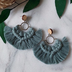 May include: A pair of handmade, teal macrame earrings. Each earring features a wooden stud, a gold-colored metal ring, and a fan-shaped design with knotted macrame and fringe. The earrings are displayed on a marble surface.