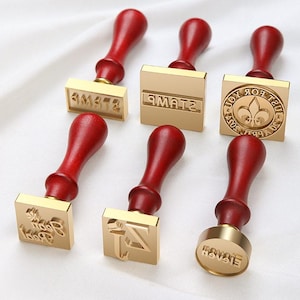 May include: A collection of six wax seal stamps with red wooden handles and gold-colored stamp heads. The stamp heads feature various designs, including text and a fleur-de-lis symbol. The stamps are arranged on a white surface.