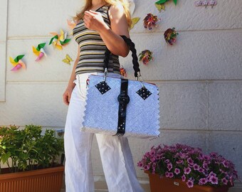 May include: A woman wearing white trousers and a striped vest top carries a white and black woven tote bag with a black buckle and silver studs. The bag has a black handle and a black and white patterned design.