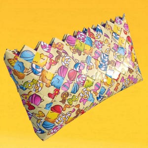May include: A woven clutch featuring a repeating pattern of colorful candies on a light yellow background. The clutch has a rectangular shape with a zig-zag top edge and a zipper closure. The candies include lollipops, ice cream cones, and other sweets.