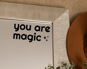 May include: A mirror with a silver frame and a white background. The mirror has a sticker on it that says "you are magic." There is a brown hat and a plant in the background.