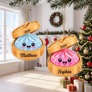 May include: Two hanging ornaments shaped like bamboo steamer baskets. One contains a blue glittery dumpling with the name Matthew and 2026 printed on it. The other contains a pink glittery dumpling with the name Sophia and 2026 printed on it.