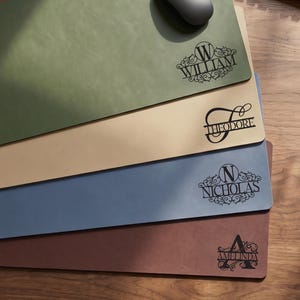May include: Four personalized desk mats in green, tan, blue, and brown. Each mat features a name in a decorative font. A computer mouse rests on the green mat. The mats are rectangular and made of a leather-like material.