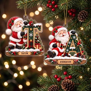 May include: Two flat Christmas ornaments featuring Santa Claus figures leaning against large letters T and A. The letters are dark green with gold stars, snowflakes, and plaid patterns. Each ornament has a nameplate reading TATUM or ANDERSON.