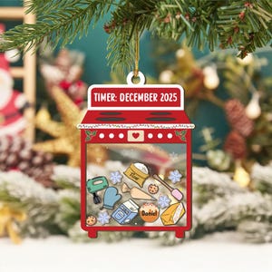 May include: A red oven-shaped Christmas ornament with the text "TIMER: DECEMBER 2025". The ornament features baking-themed illustrations inside, including flour, milk, and a mixer. It is hanging from a gold string against a blurred background.