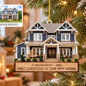 May include: A house-shaped Christmas ornament featuring a dark blue and white two-story home with a front porch. A wooden base displays the text 21 WILLOW GROVE - 2026 FIRST CHRISTMAS IN OUR NEW HOME. The ornament hangs from a gold string.