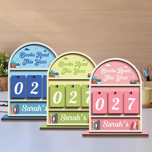 May include: Three wooden book counters in blue, green, and pink, each with the text "Books Read This Year." The counters display numbers, with "02" and "27" visible. Each counter has a shelf with books and the name "Sarah's."