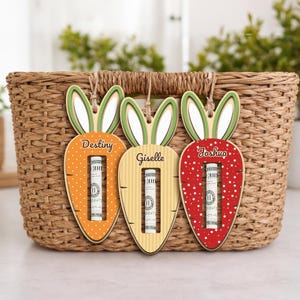 May include: Three wooden carrot-shaped Easter gift holders with bunny ears. Each carrot has a name: Destiny, Giselle, and Joshua. The carrots are orange, yellow, and red, with money visible in the center. They hang in front of a woven basket.