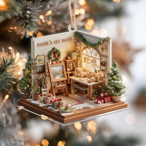 May include: A Christmas ornament shaped like a miniature art studio. The ornament features a small easel with a painting, a table with gifts, and a Christmas tree. The text "Sophia's Art Studio" is visible on the ornament.