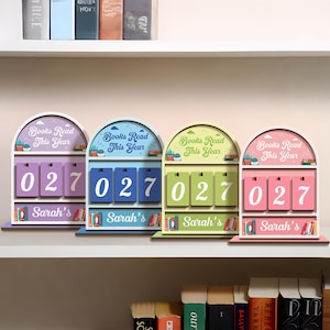 May include: Four colorful wooden book counters with the text "Books Read This Year" and the number "027". Each counter has a different color scheme: purple, blue, green, and pink. The name "Sarah's" is printed on the bottom.