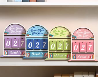 May include: Four colorful wooden book counters with the text "Books Read This Year" and the number "027". Each counter has a different color scheme: purple, blue, green, and pink. The name "Sarah's" is printed on the bottom.